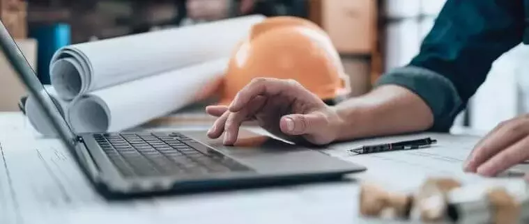 An architect working on their laptop, alongside the laptop are rolled up plans.